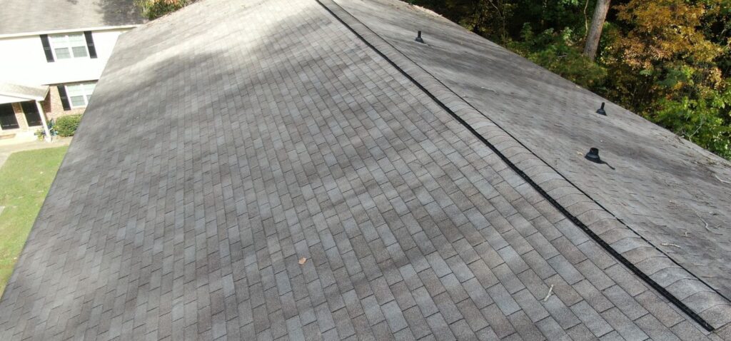A residential roof with gray 3-tab asphalt shingles and a ridge vent, showing even shingle alignment and light debris near surrounding trees.
