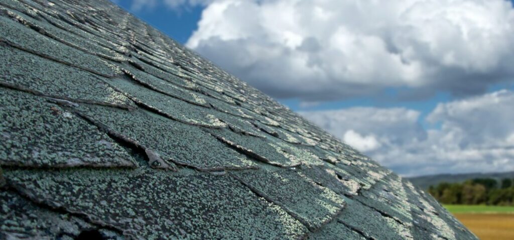 Close-up of an aging asphalt shingle roof with visible wear and granule loss under a cloudy sky.